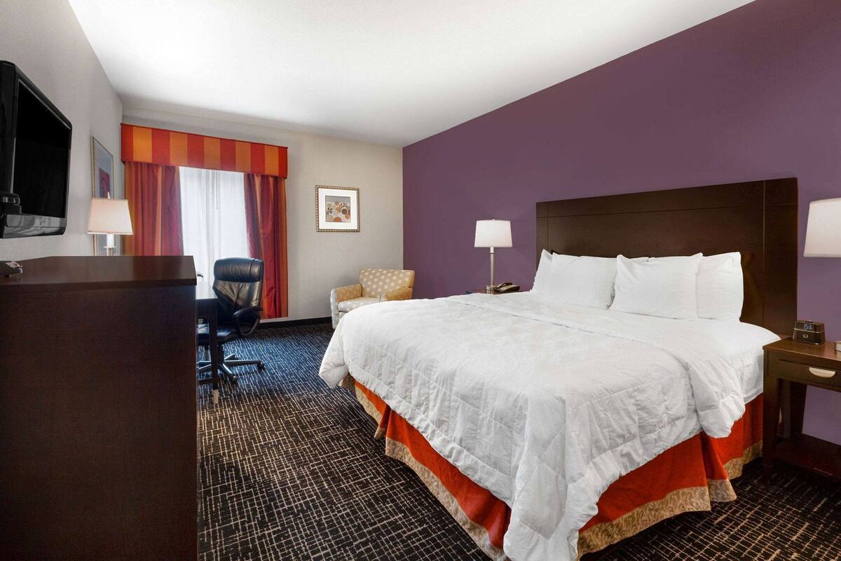 A hotel room with a large bed, a desk, a chair, a TV, and a window with curtains.