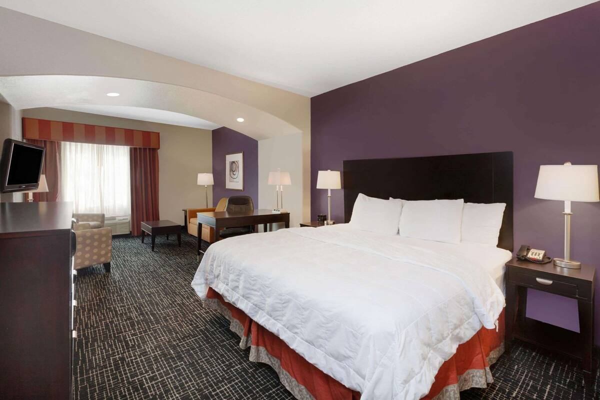 A hotel room with a large bed, a desk, a chair, a TV, and a desk lamp.