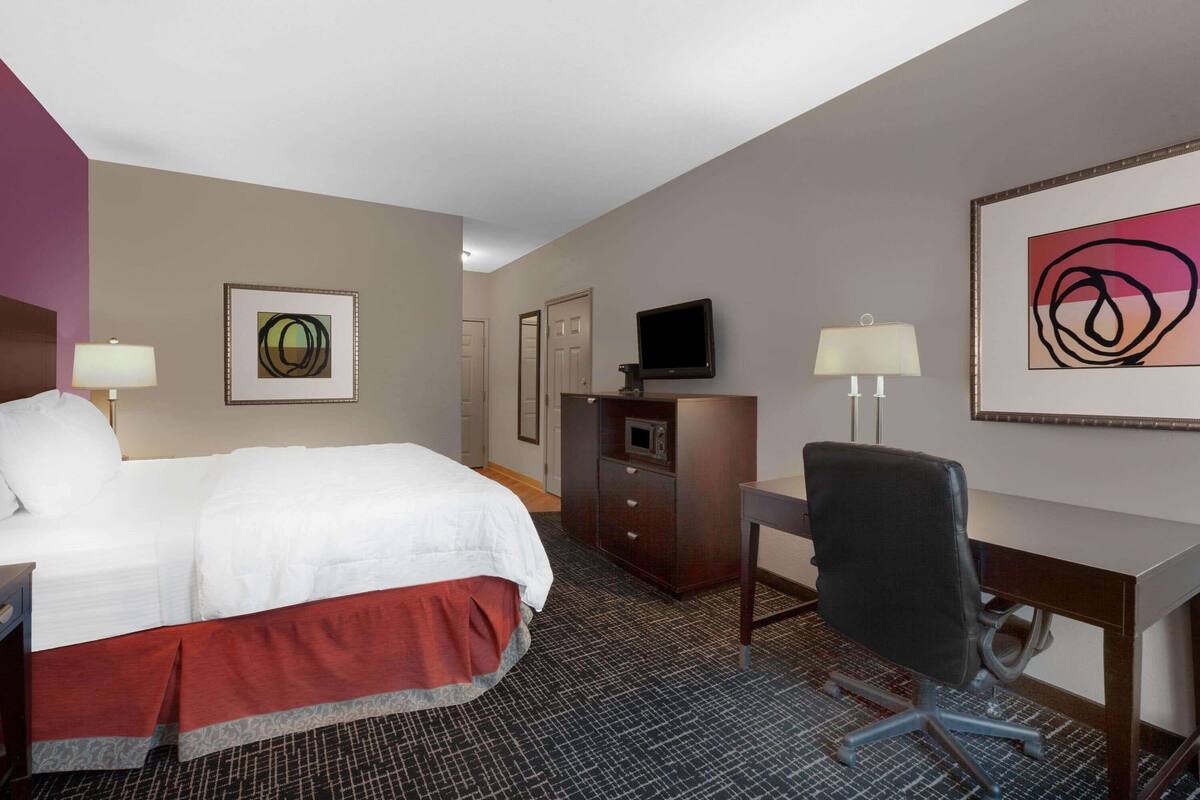 A hotel room with a bed, desk, chair, TV, and artwork on the walls.