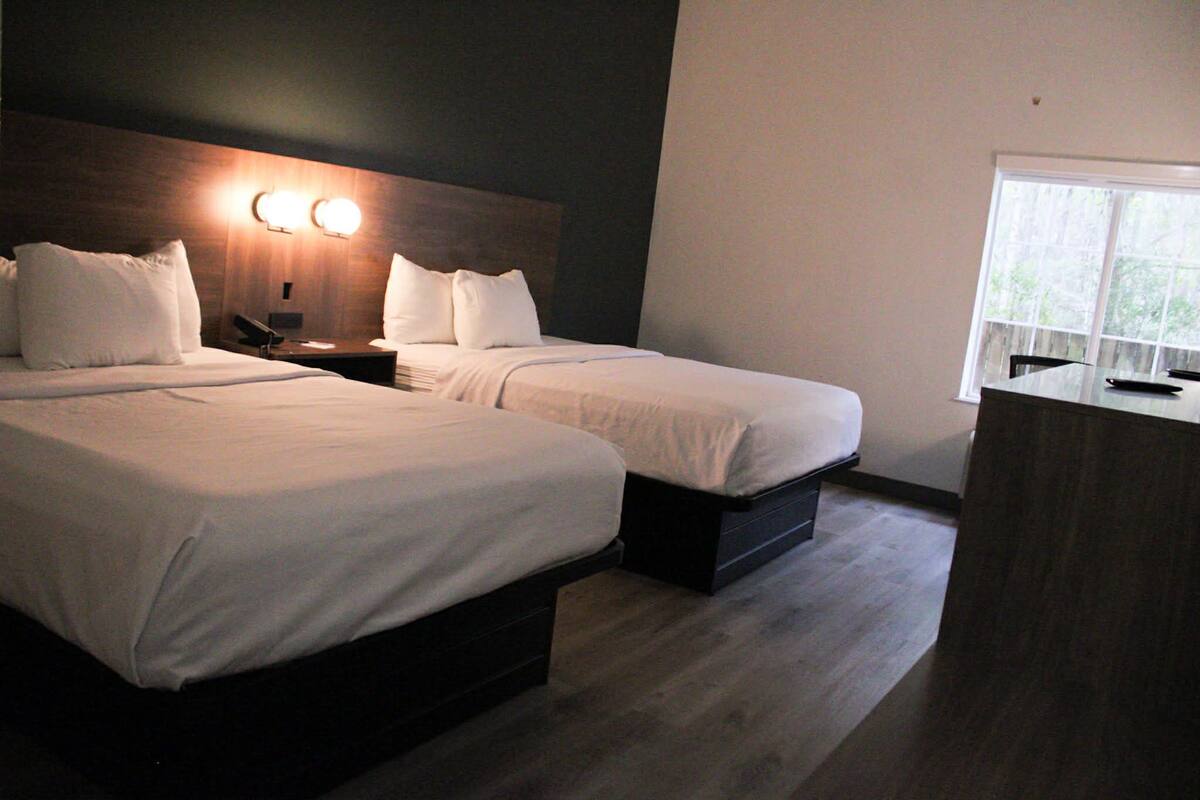 A hotel room with two single beds, a nightstand with a lamp, a desk with a chair, and a large window.