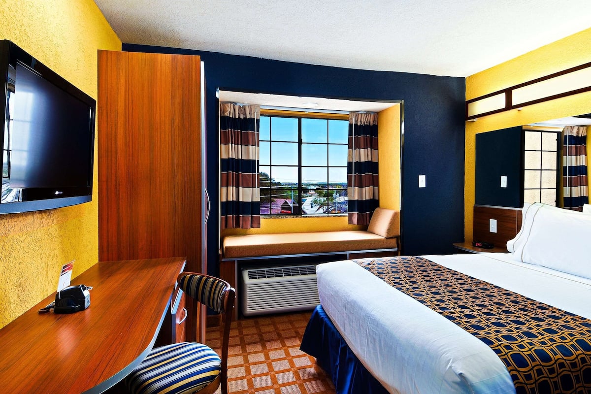 A hotel room with a large bed, a desk with a chair, a television, a window with curtains, and a view of the outside.