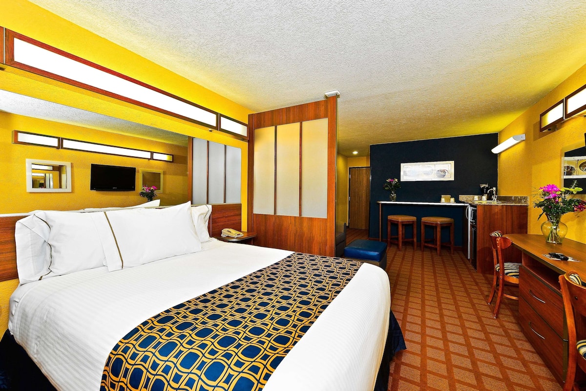 A hotel room with a large bed, a desk with two chairs, a small kitchenette, and a bathroom with a mirror and sink.