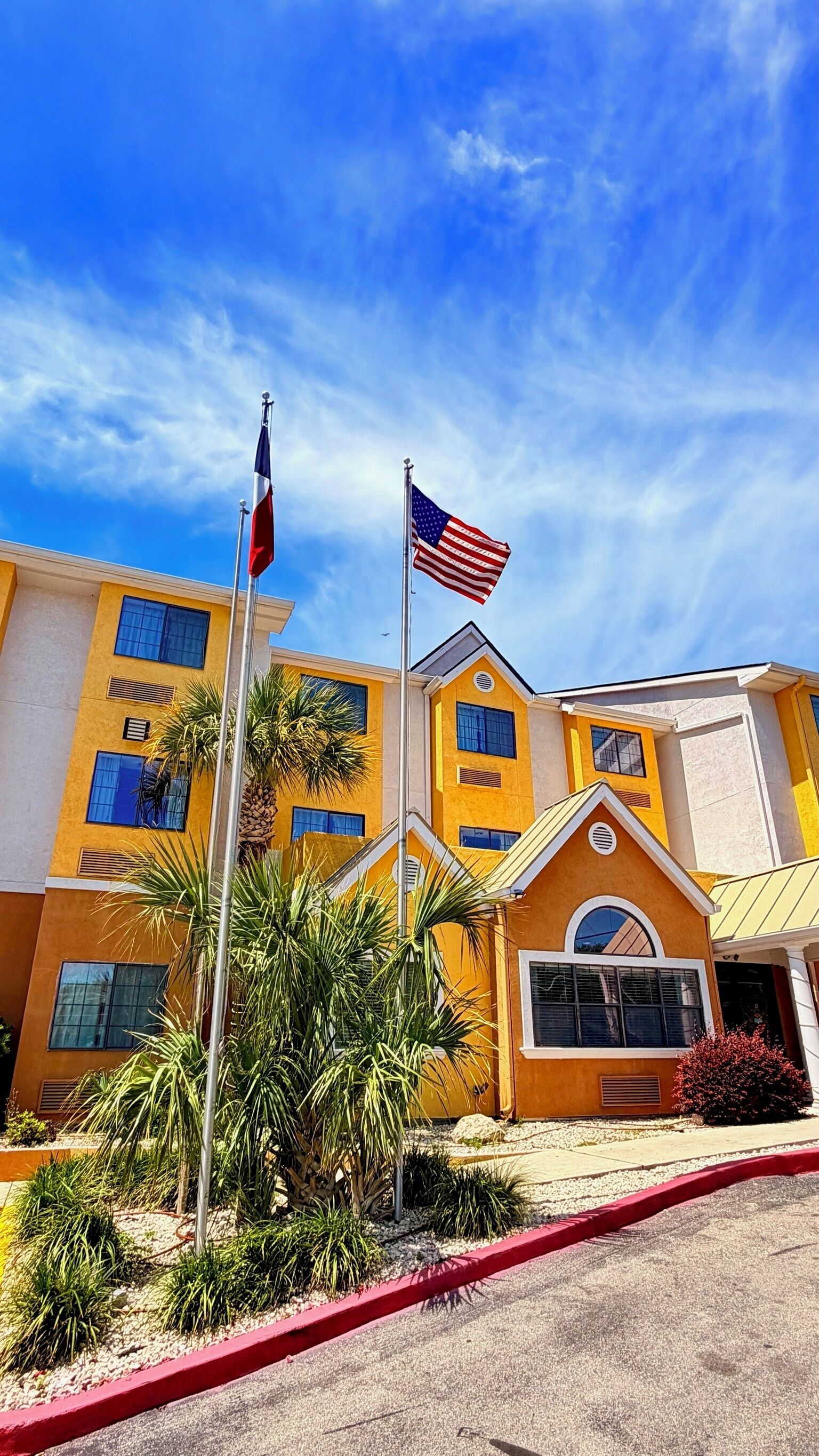 Un edificio amarillo con dos banderas, palmeras y una bordillo rojo.