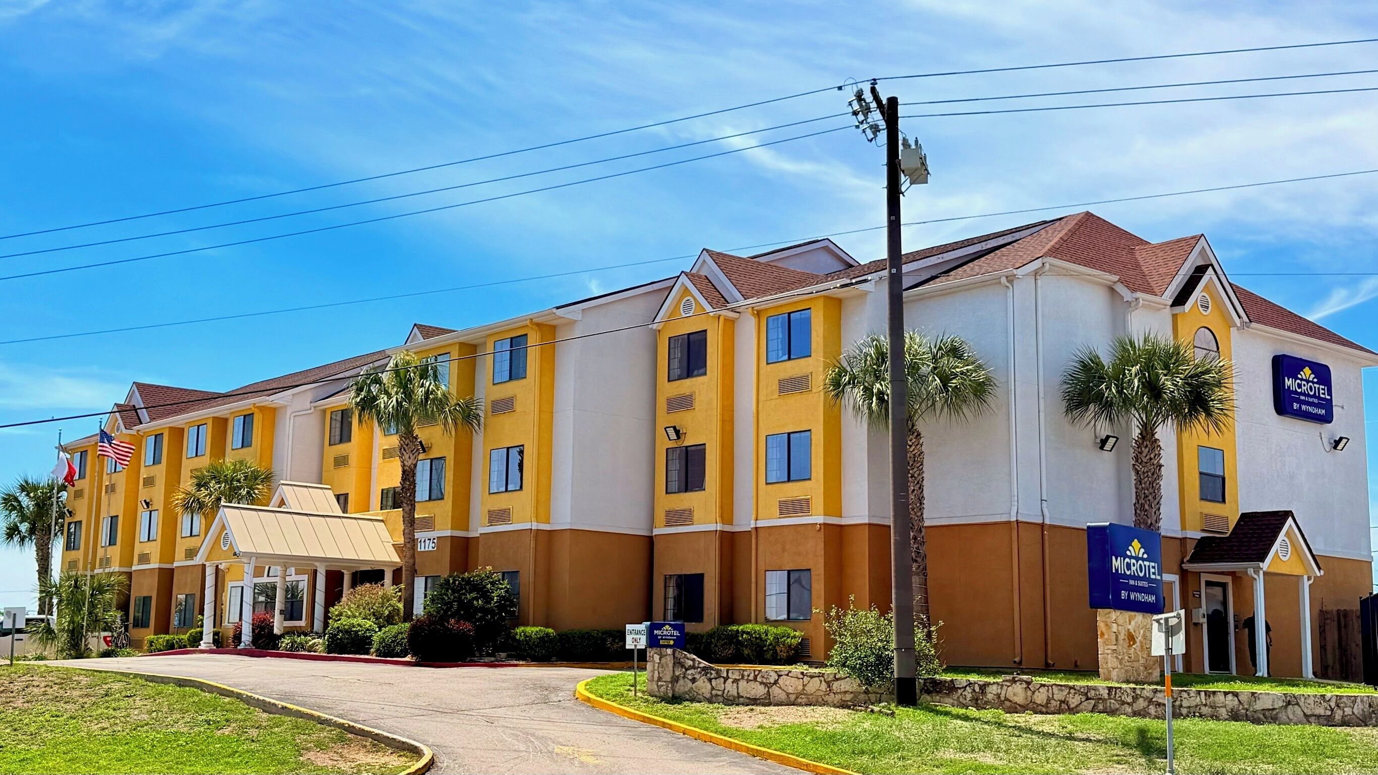 Un hotel de varios pisos con fachada amarilla y blanca, palmeras y un cielo azul despejado.