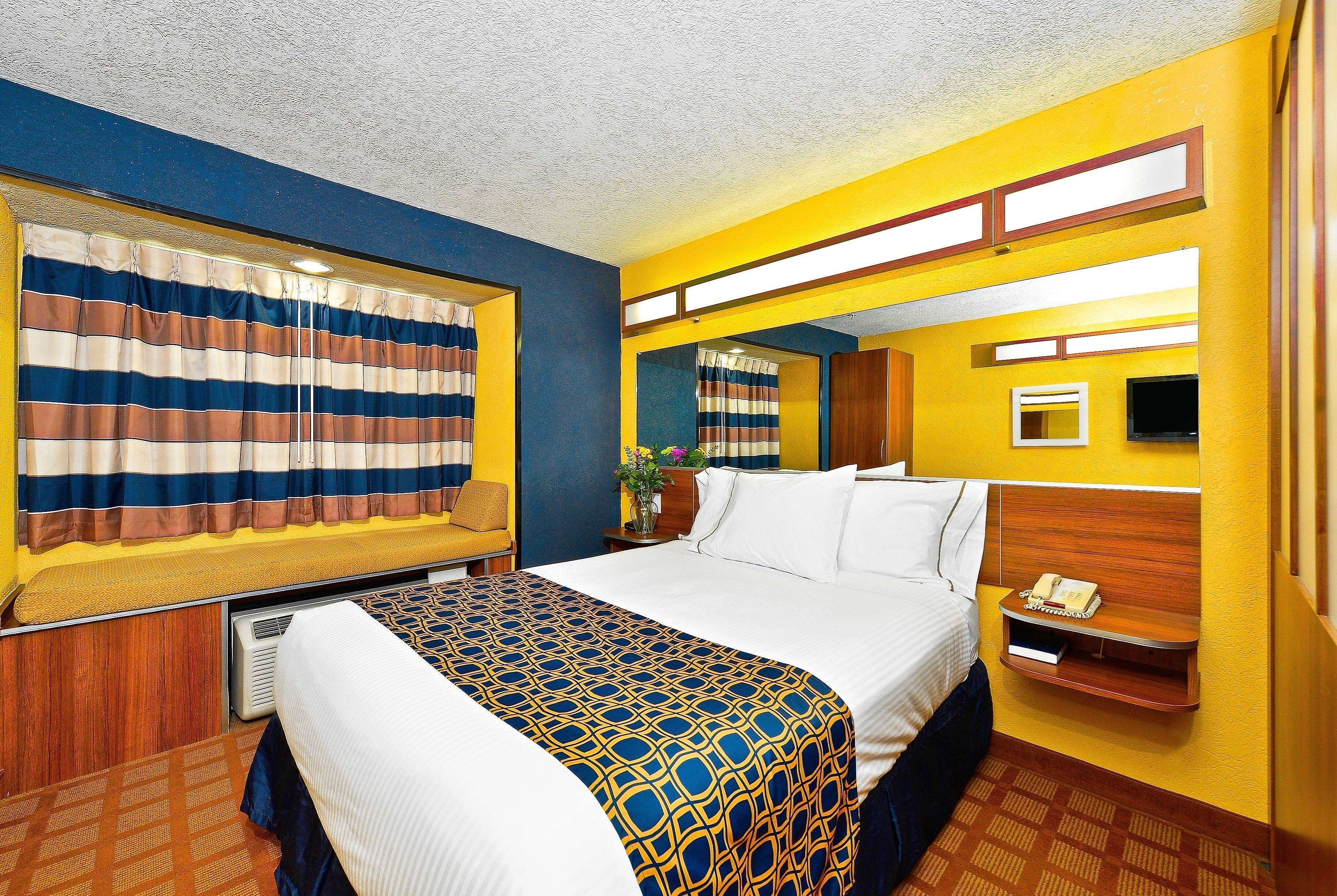 Une chambre d’hôtel avec un lit, une table de chevet avec un téléphone, une fenêtre avec des rideaux à rayures et un miroir au mur.