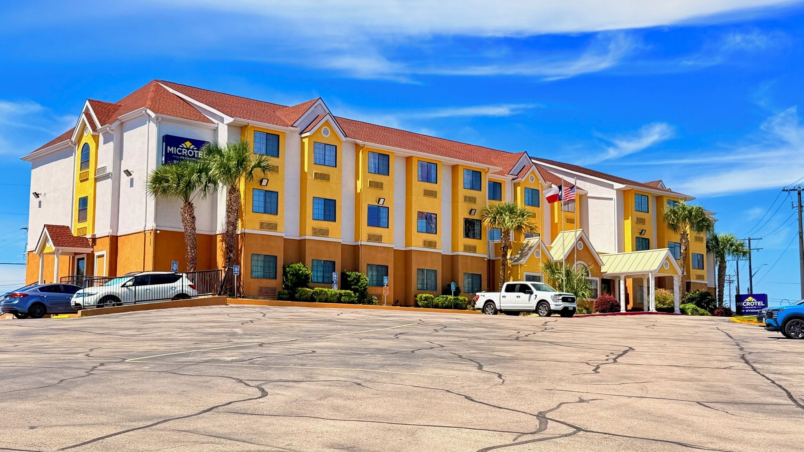 Un hotel a più piani con facciata gialla, palme e un parcheggio.