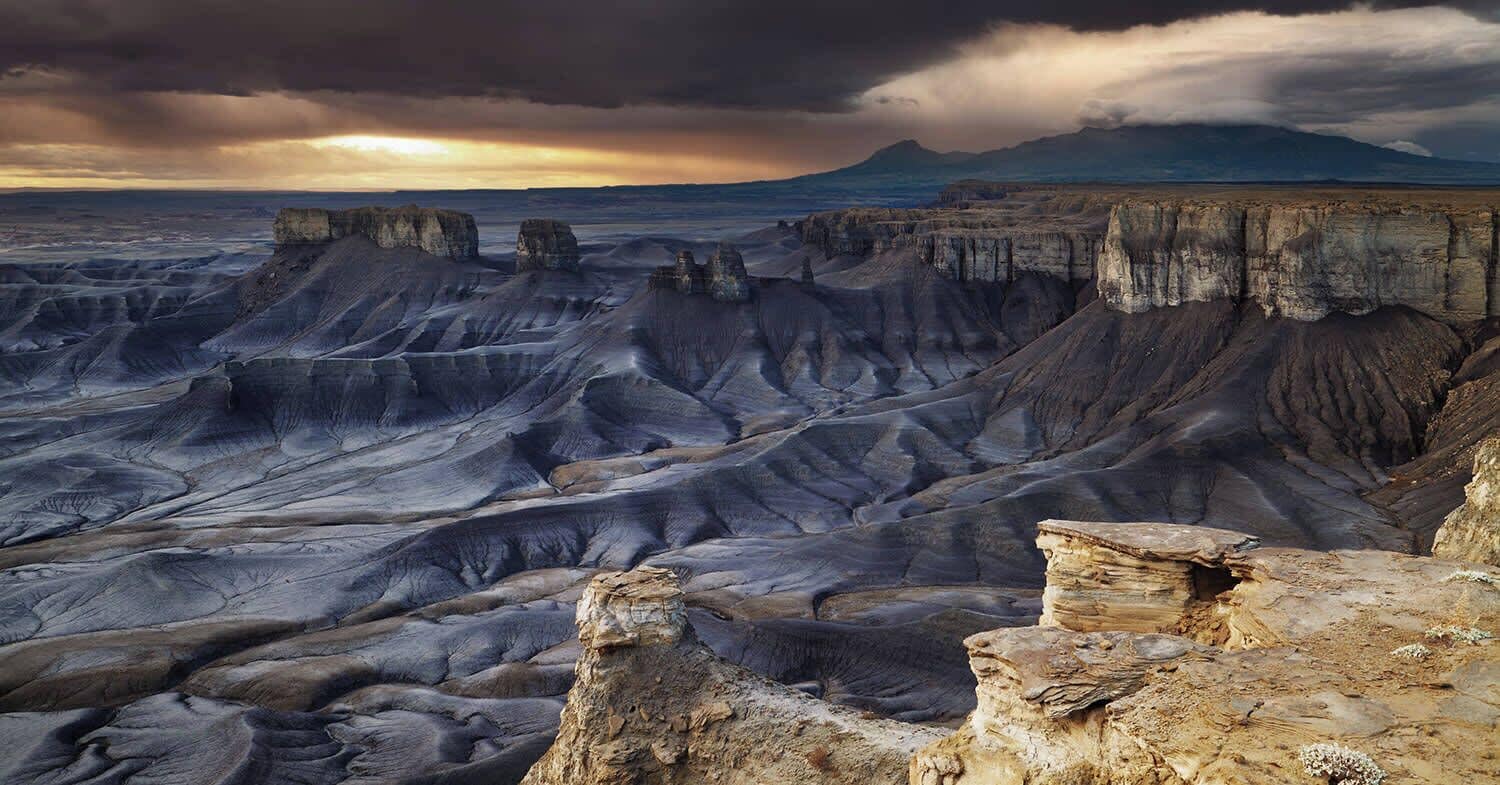 Une vue panoramique d’un paysage accidenté, avec des formations rocheuses imposantes et une chaîne de montagnes lointaine, le tout sous un ciel spectaculaire.