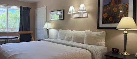Habitación de hotel con una cama grande, dos lámparas de noche, una mesita de noche, un escritorio, una silla, una ventana con cortinas y un cuadro enmarcado en la pared.