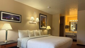 Habitación de hotel con una cama grande, dos lámparas de noche, una mesita de noche, un cuadro enmarcado con un paisaje, un espejo de baño iluminado y una lámpara de techo.