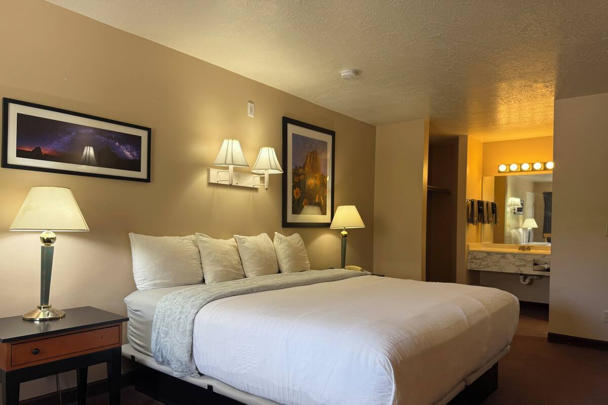 Une chambre d’hôtel avec un grand lit, deux lampes de chevet, une table de chevet, un tableau représentant un paysage, un miroir avec éclairage dans la salle de bain et un luminaire au plafond.