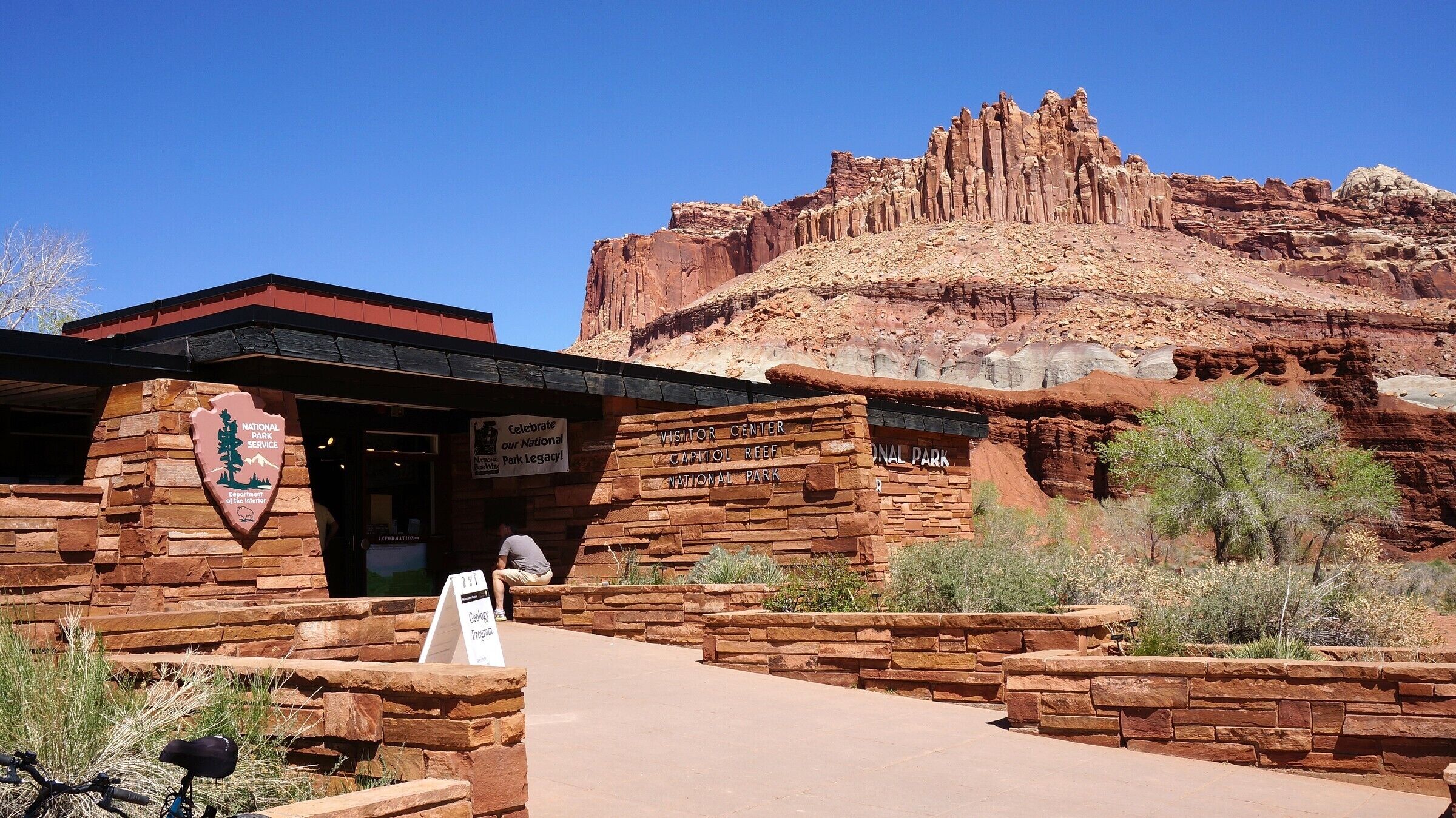 A visitor center with a stone facade, a sign for the Geology Program, and a backdrop of red rock formations.