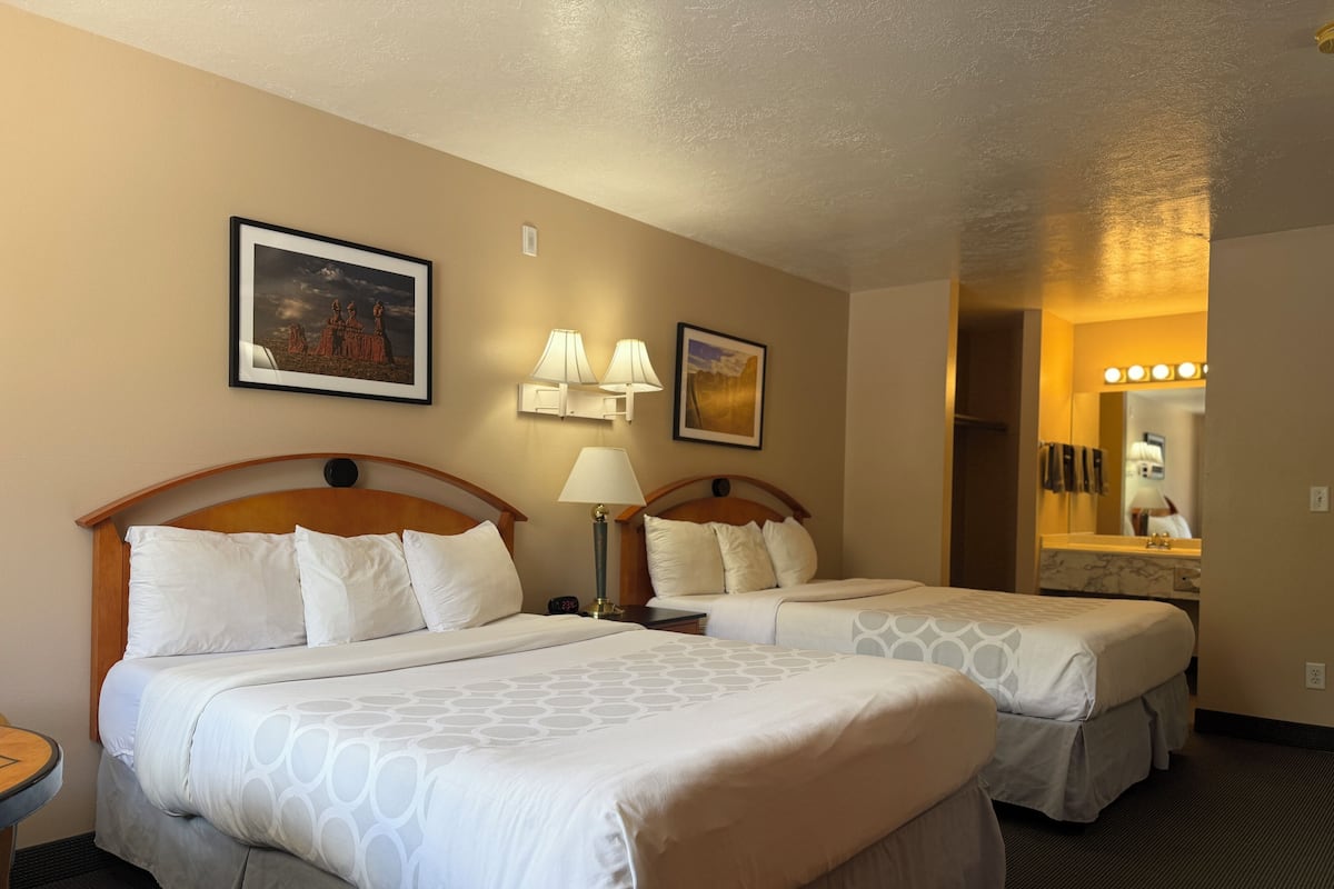 Une chambre d’hôtel avec deux lits, une petite table et une chaise. Des lampes murales et un tableau encadré sont accrochés au mur, au-dessus des lits.