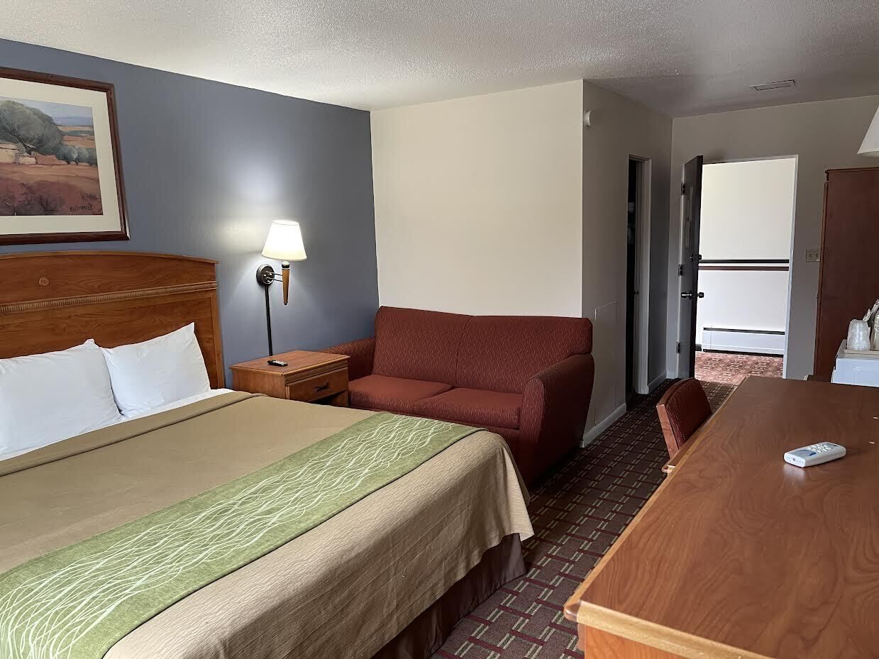 A hotel room with a bed, a red sofa, a wooden table, and a lamp.