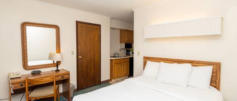 Habitación de hotel con cama, escritorio con silla, espejo, puerta de madera y una kitchenette al fondo.
