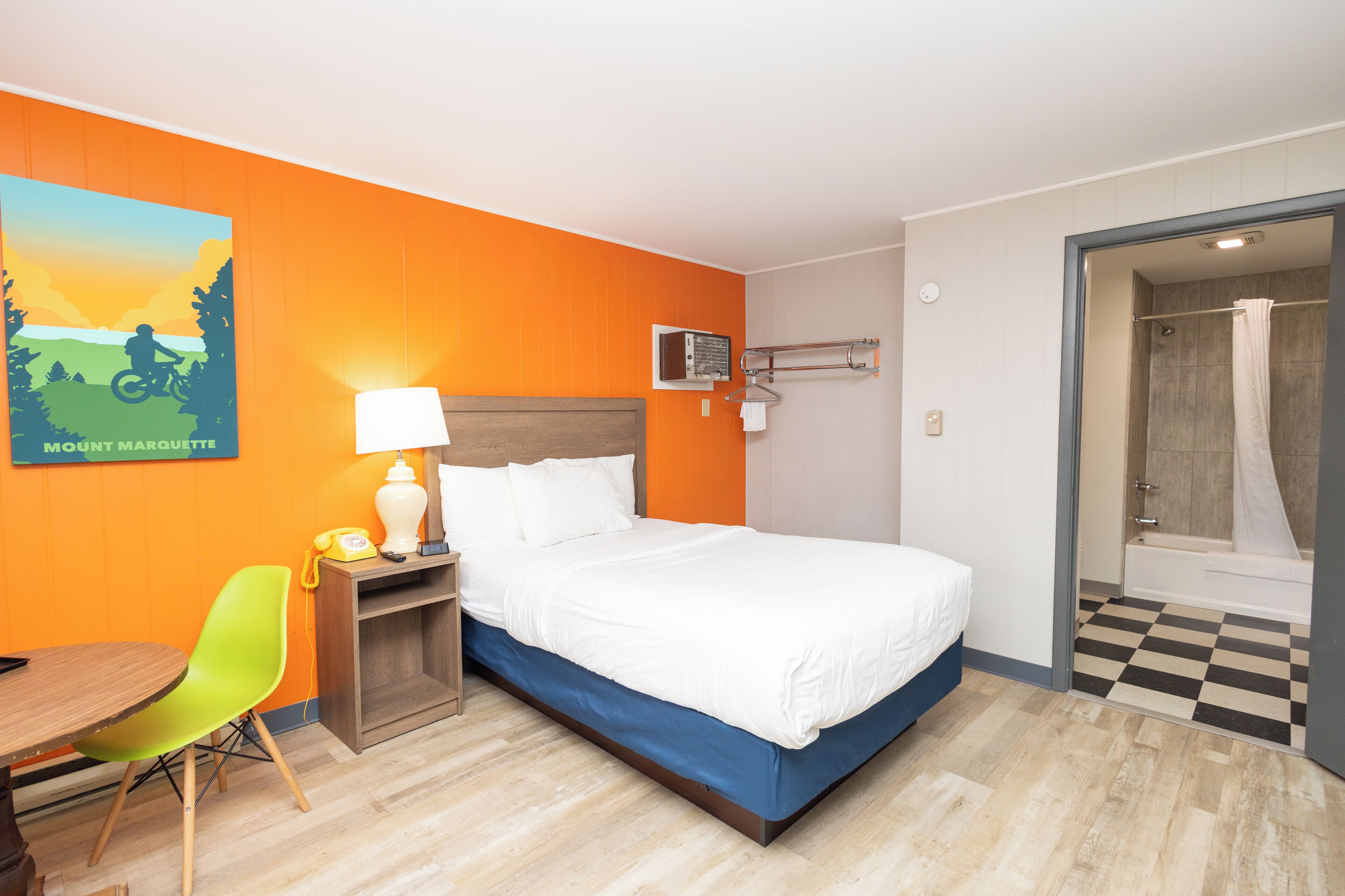 A hotel room with a bed, a desk with a chair, a shower, and a wall-mounted radio.