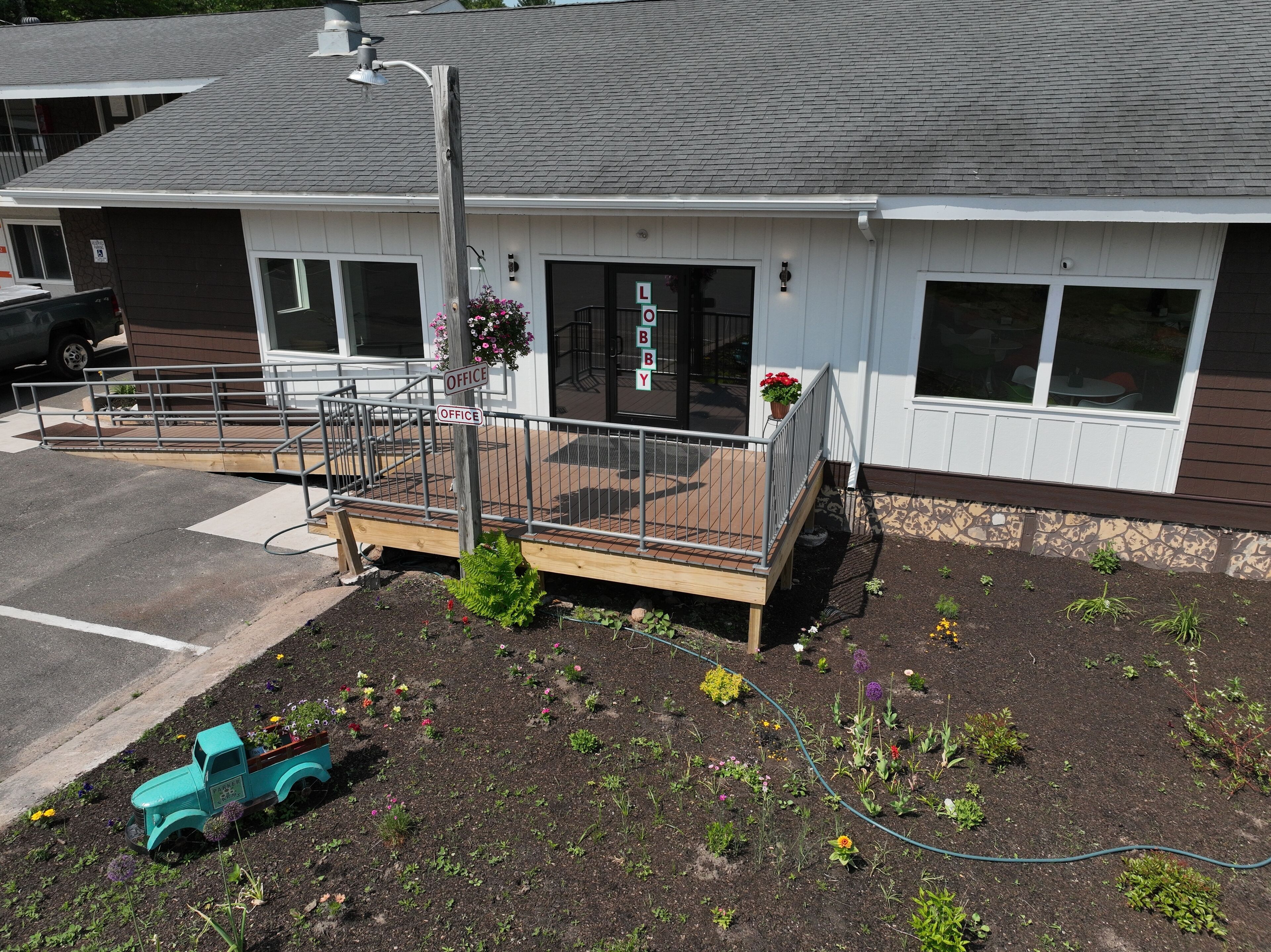 A building with a white exterior, a covered porch with a metal railing, and a small garden area with various plants and flowers.