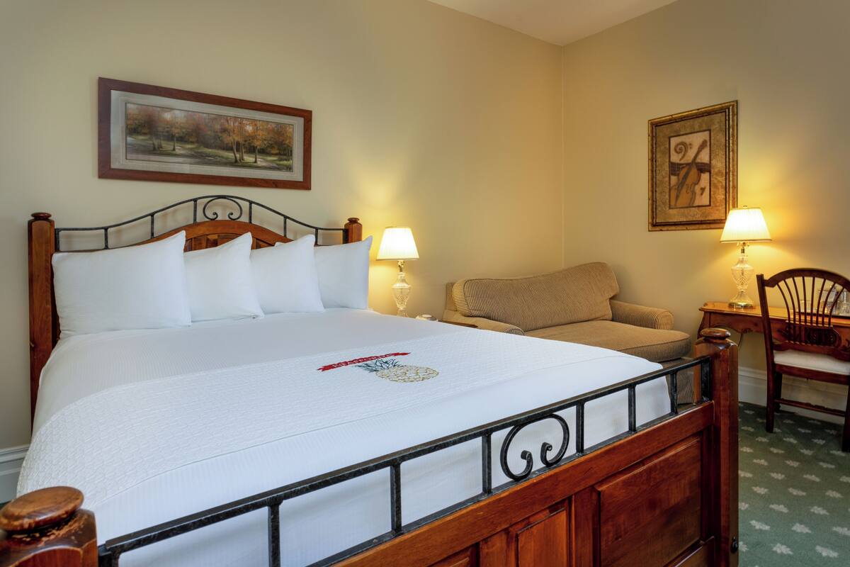 A neatly made bed with a white comforter and a pineapple design, a wooden headboard, two bedside lamps, a chair, and a framed picture on the wall.