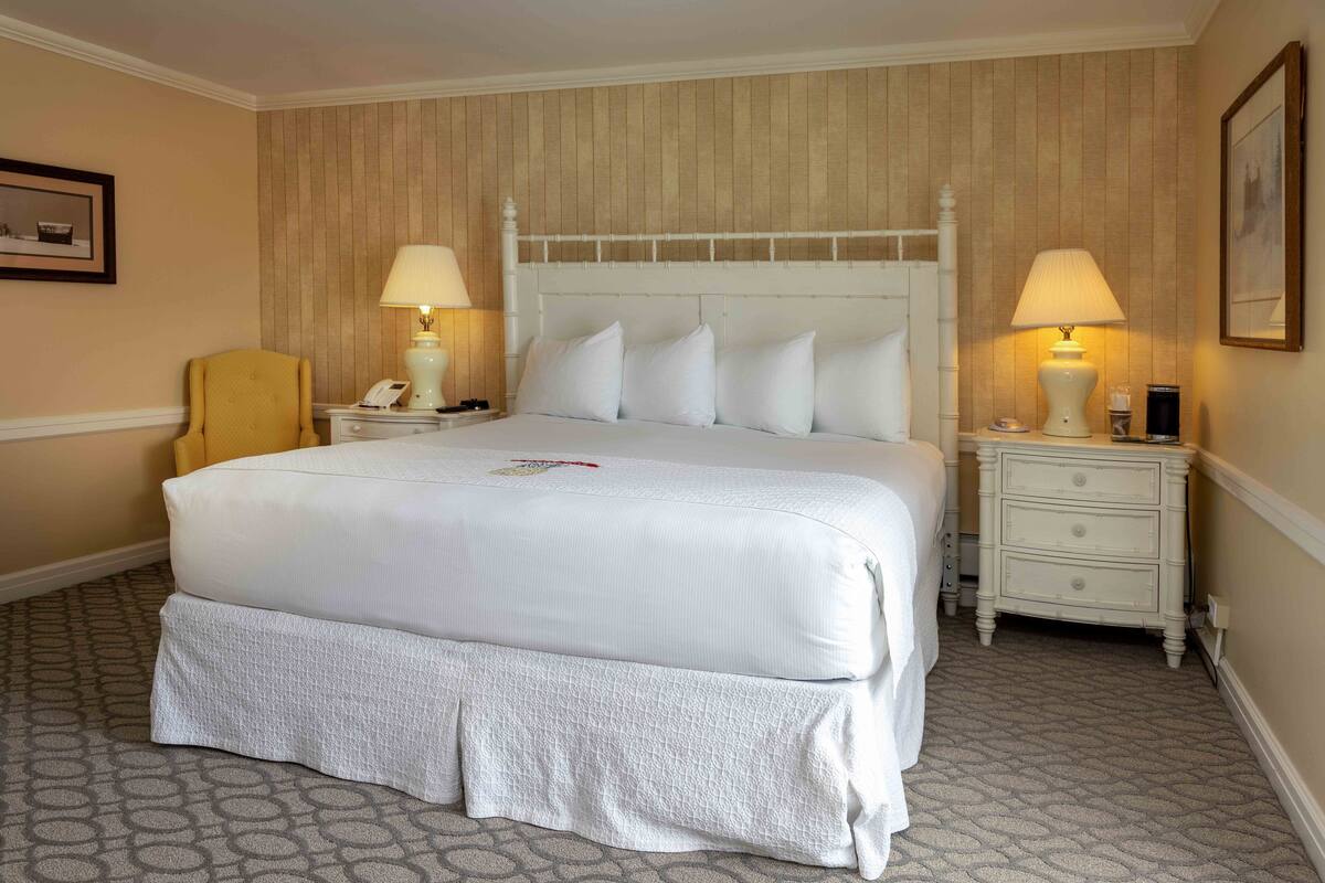 A neatly made bed with white bedding, two bedside tables with lamps, a chair, and a patterned carpet.