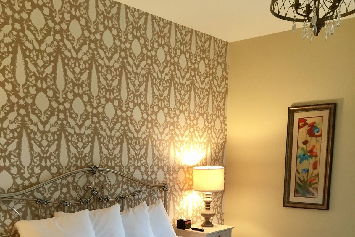 A neatly made bed with white linens and a decorative pillow, a bedside table with a lamp, and a patterned wallpaper.