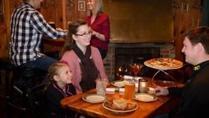 A group of people seated around a wooden table with a pizza and drinks, in a room with wooden walls and a fireplace. - Hotel Perry (Petoskey)