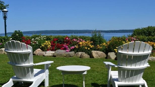Two white Adirondack chairs and a small round table on a grassy lawn with a view of a body of water and hills in the distance. - Hotel Perry (Petoskey)