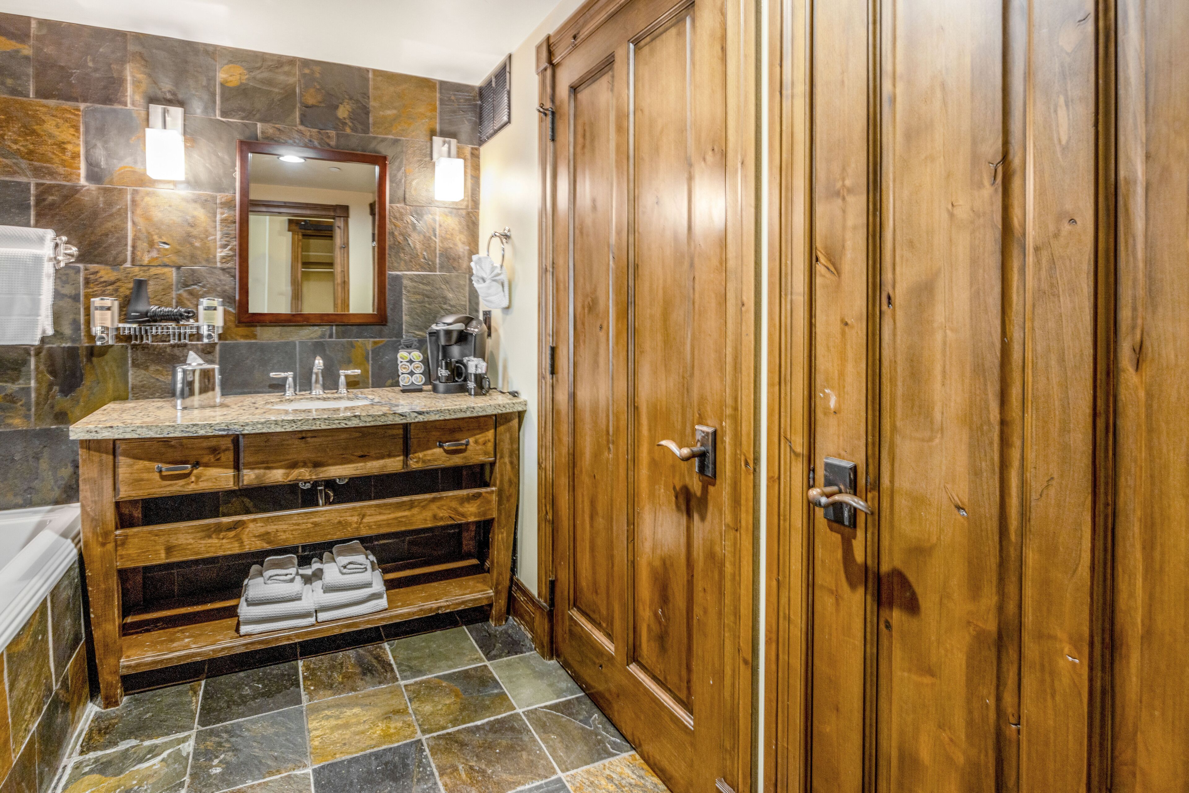 A bathroom with a wooden vanity, a large mirror, and a stone-tiled floor.