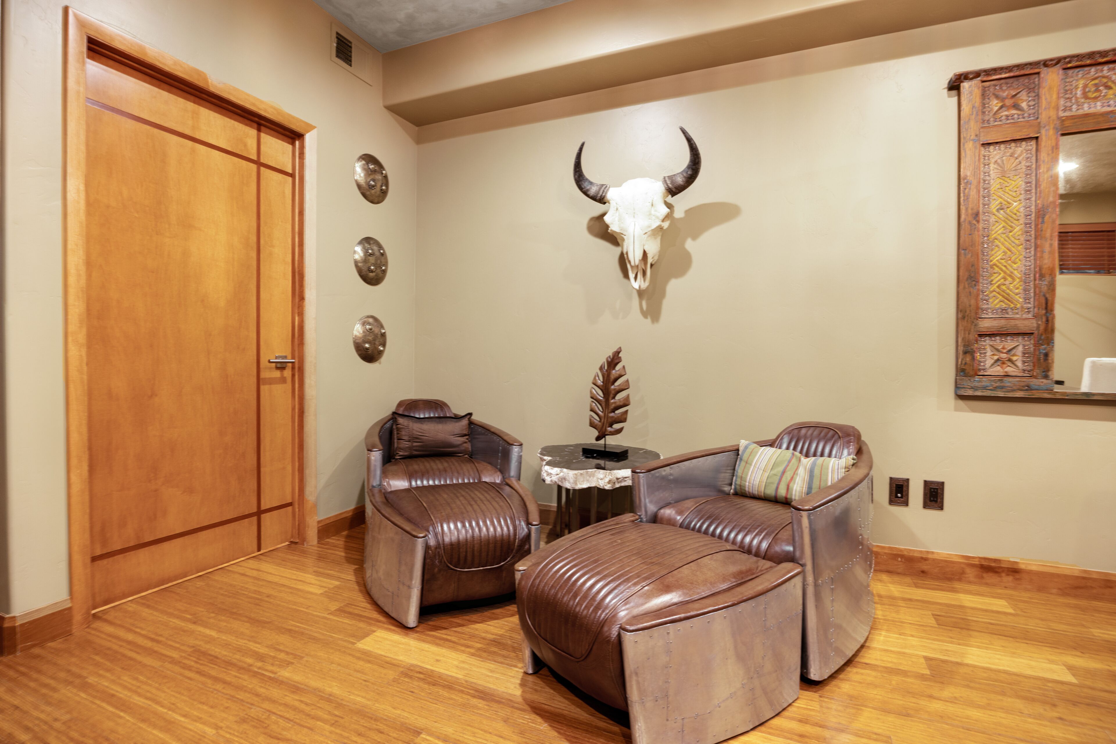 A room with a wooden door, two leather chairs, a small table with a decorative item, and a wall-mounted bull skull.