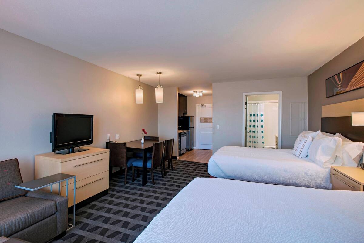 A hotel room with two beds, a desk with a TV, a sofa, and a kitchenette in the background.