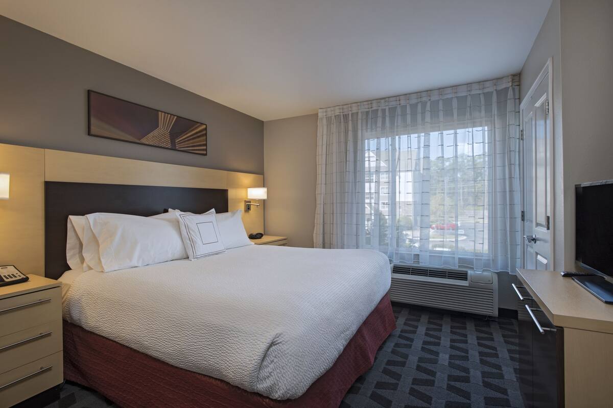 A hotel room with a large bed, a desk with a computer, a TV, and a window with curtains.
