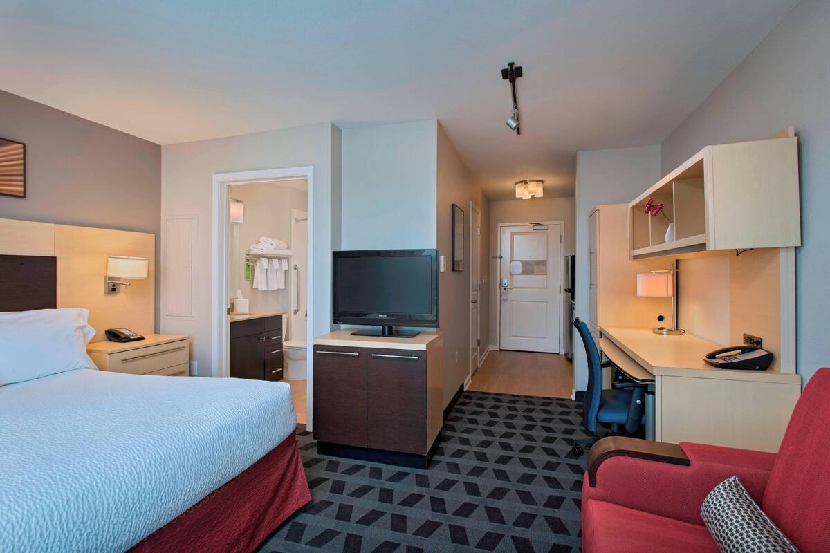 A hotel room with a bed, a desk, a chair, a television, and a small kitchenette.