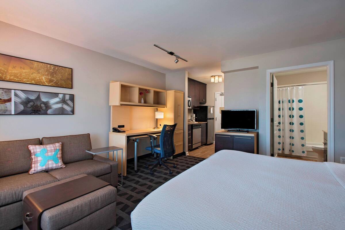 A hotel room with a bed, a desk, a chair, a sofa, a TV, a kitchenette, and artwork on the walls.