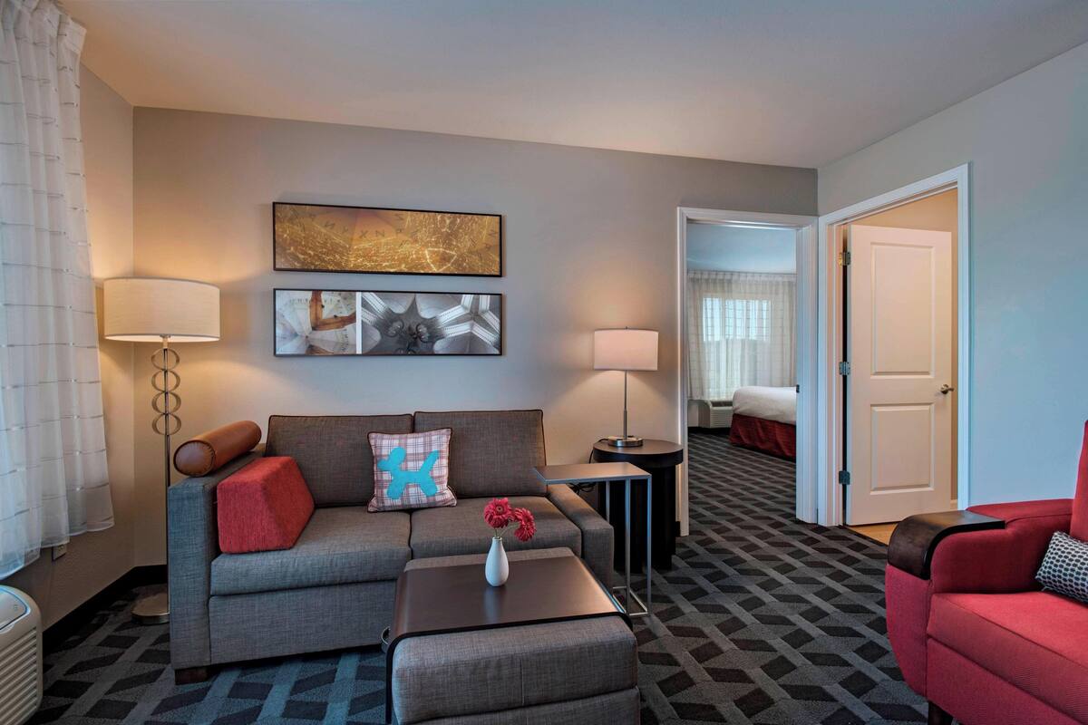 A hotel room with a grey sofa, a red armchair, a small table with a vase of flowers, and a patterned carpet.