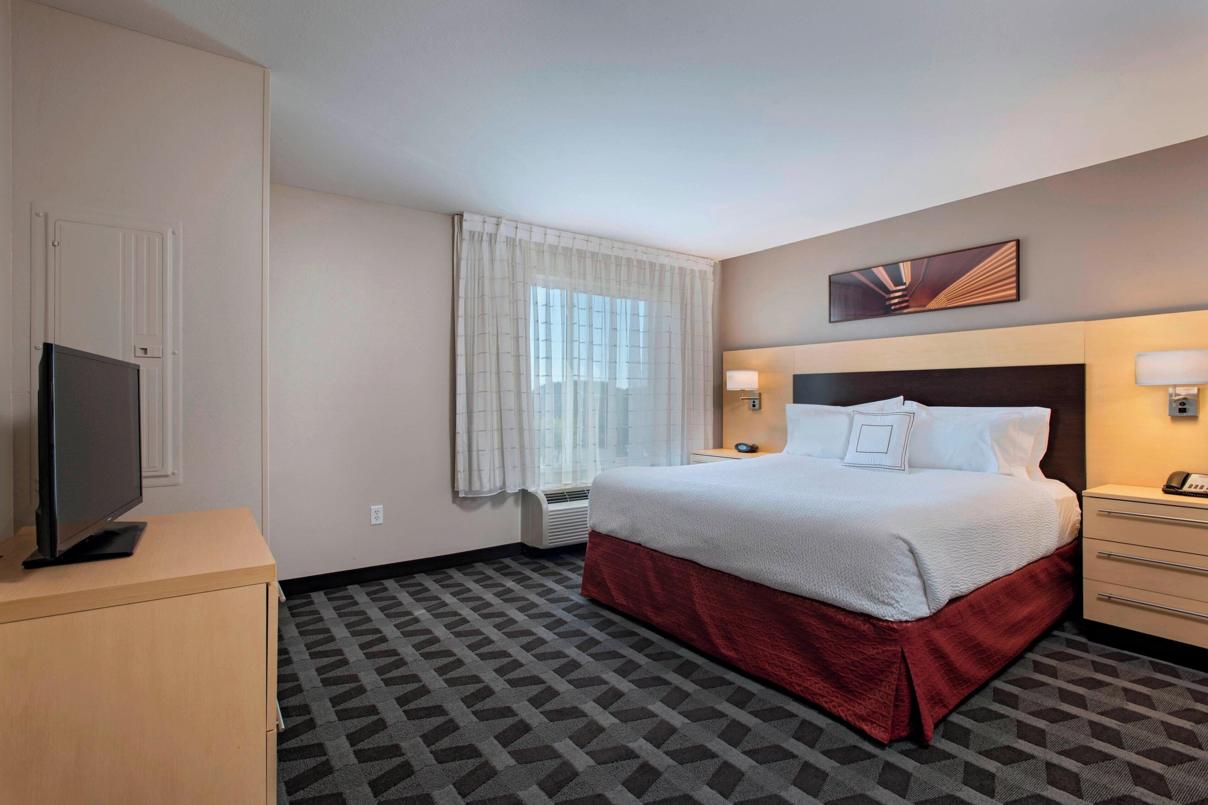 A hotel room with a bed, a TV, a desk, a lamp, and a window with curtains.