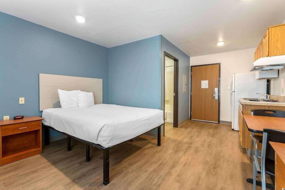 A room with a bed, a desk, a chair, a nightstand, and a door leading to a bathroom.