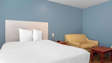 Una habitación de hotel con una cama grande, un sillón color beige, una mesita de noche de madera y una pequeña mesa.