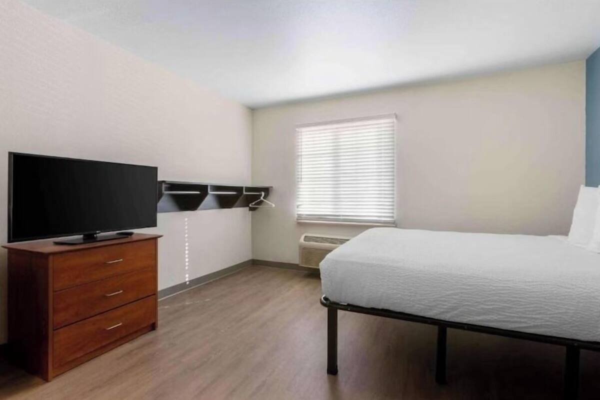 A bedroom with a bed, a TV on a wooden dresser, a window with blinds, and a wall-mounted heater.