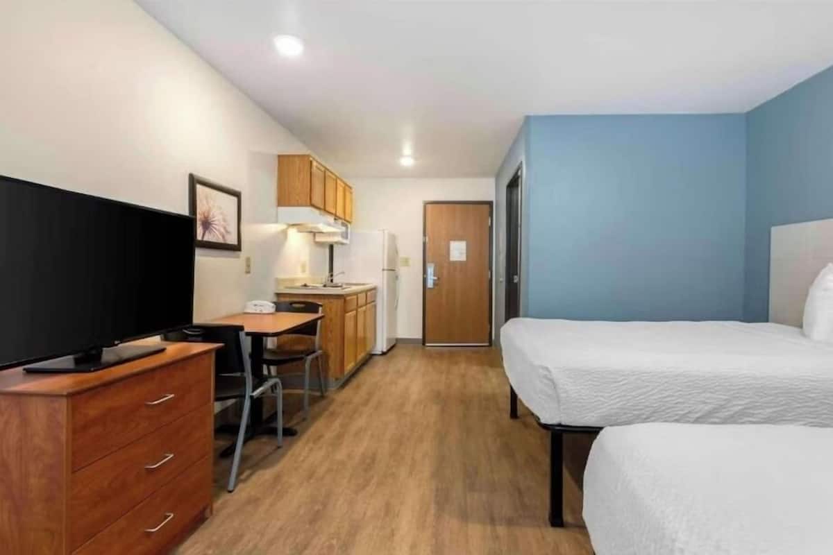 A compact hotel room with a bed, a desk with a chair, a television, a kitchenette, and a door leading to another room.