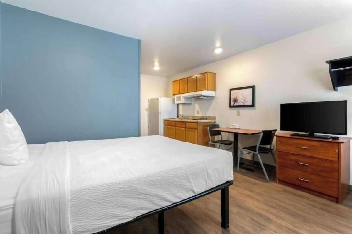 A hotel room with a bed, a desk, a chair, a television, and a kitchenette.