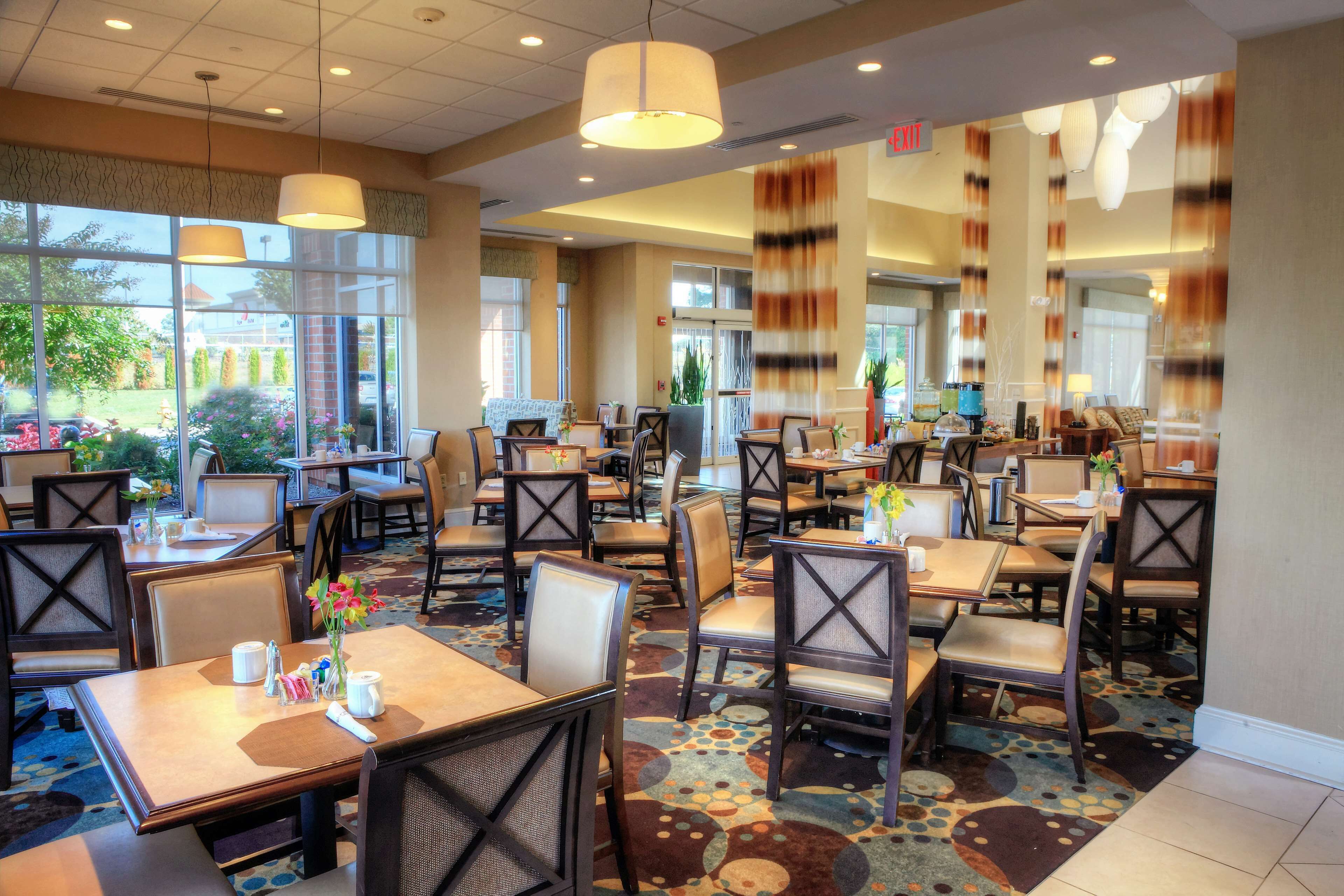 A hotel dining area with multiple tables and chairs, large windows, and a patterned carpet.