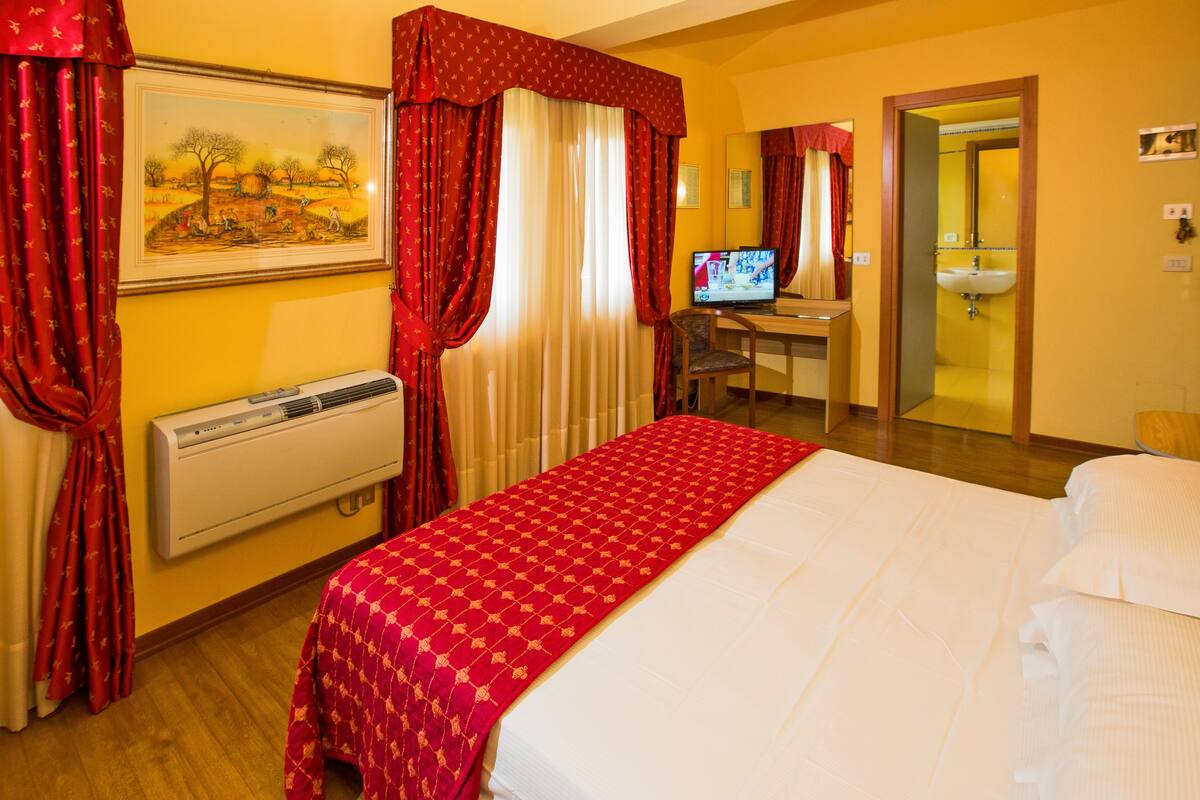 A hotel room with a bed, a desk with a computer, a TV, a bathroom, and a painting on the wall.