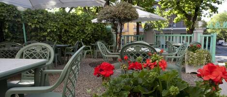 A patio area with green metal chairs and tables, surrounded by greenery and red flowers.
