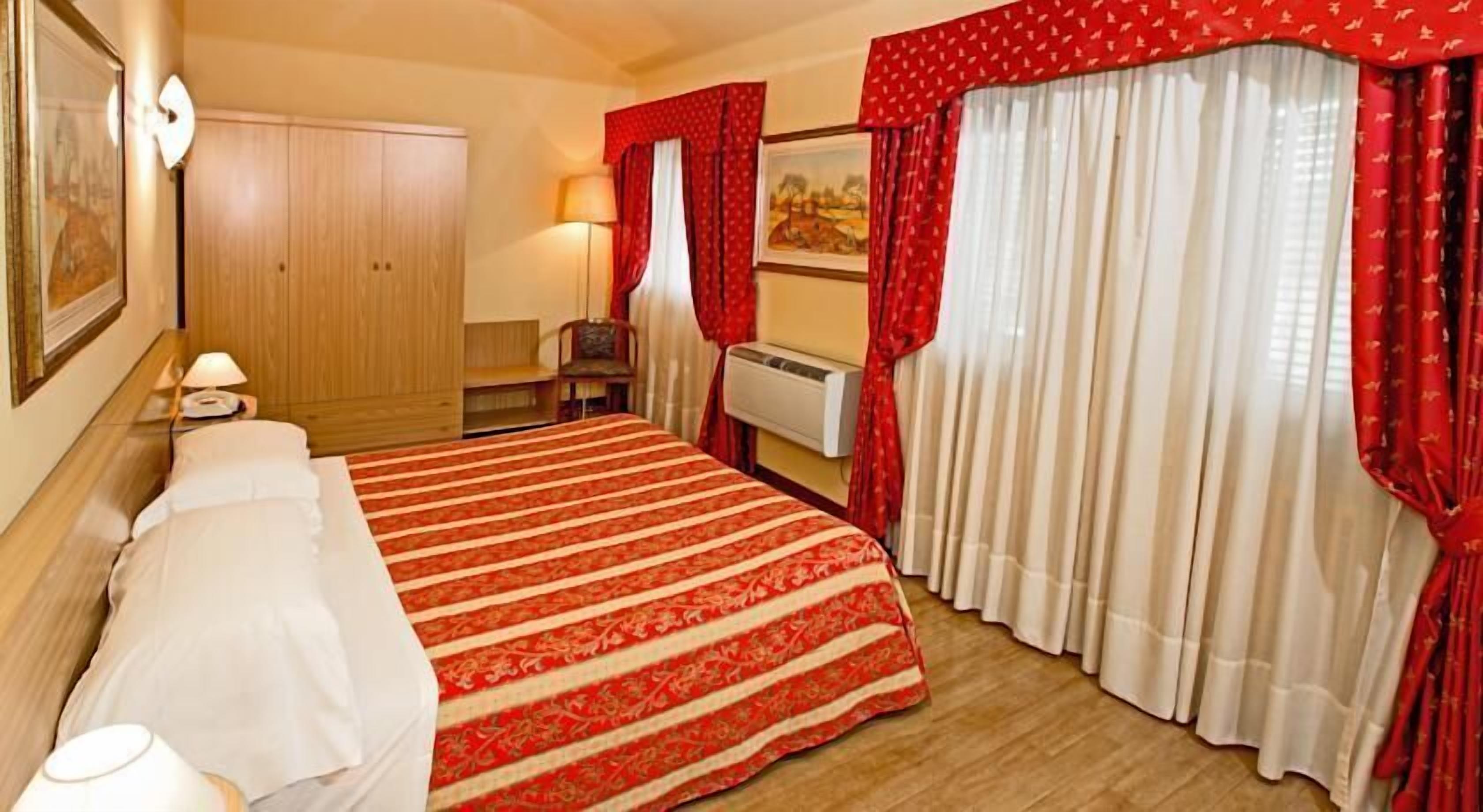 Ein Hotelzimmer mit einem großen Bett, einem Holzschrank, einem Schreibtisch mit Stuhl und einem Fenster mit roten Vorhängen.