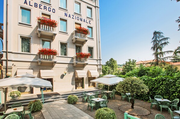 Un edificio con balcones decorados con flores rojas, mobiliario exterior con sombrillas y un jardín bien cuidado.