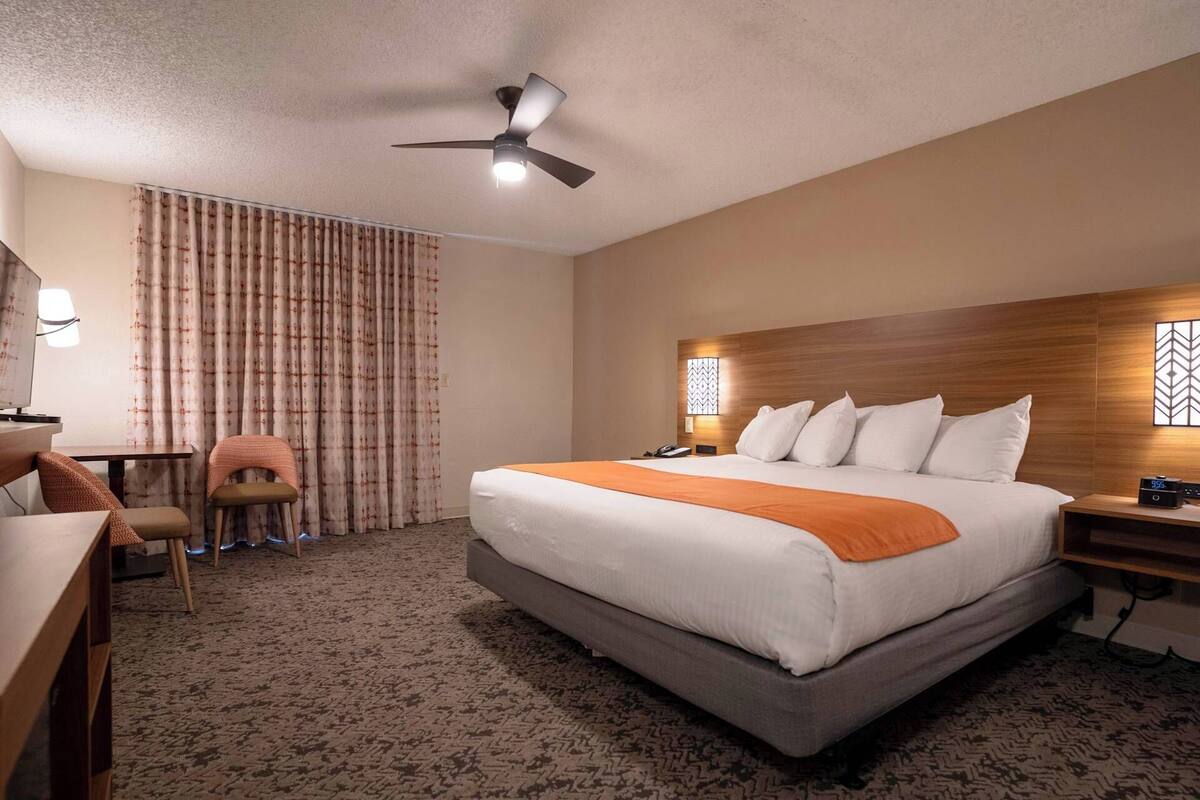 Ein Hotelzimmer mit Bett, Schreibtisch, Stuhl, Deckenventilator und Nachttisch mit einer Uhr.