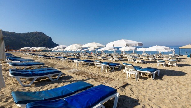 A beach with rows of sun loungers and umbrellas, a clear sky, and a mountain in the background.