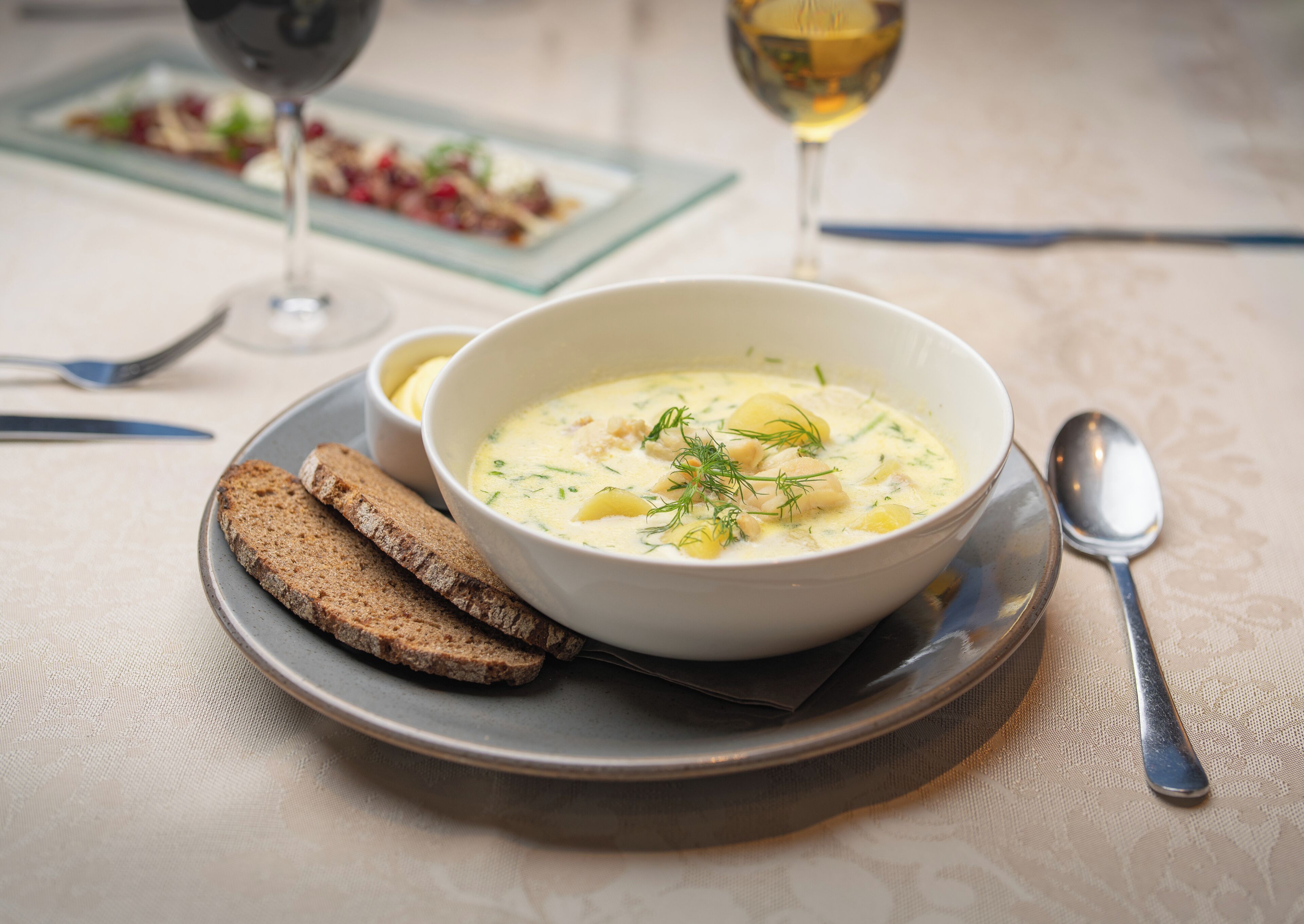 Un bol de soupe, accompagné de pain et d’une cuillère sur une assiette, avec un verre de vin blanc et une fourchette en arrière-plan.