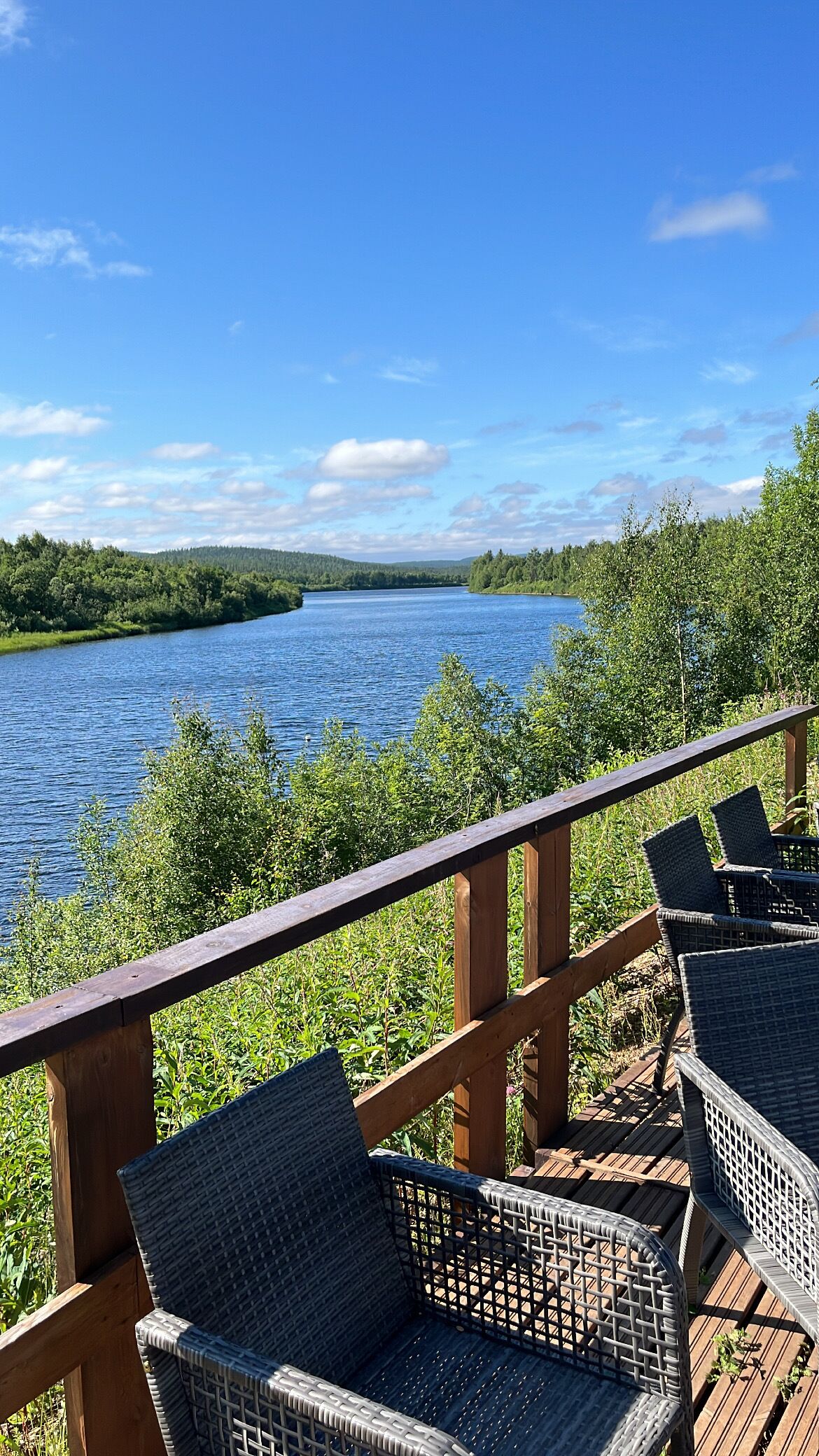 A deck with wicker chairs overlooking a lake, surrounded by greenery.