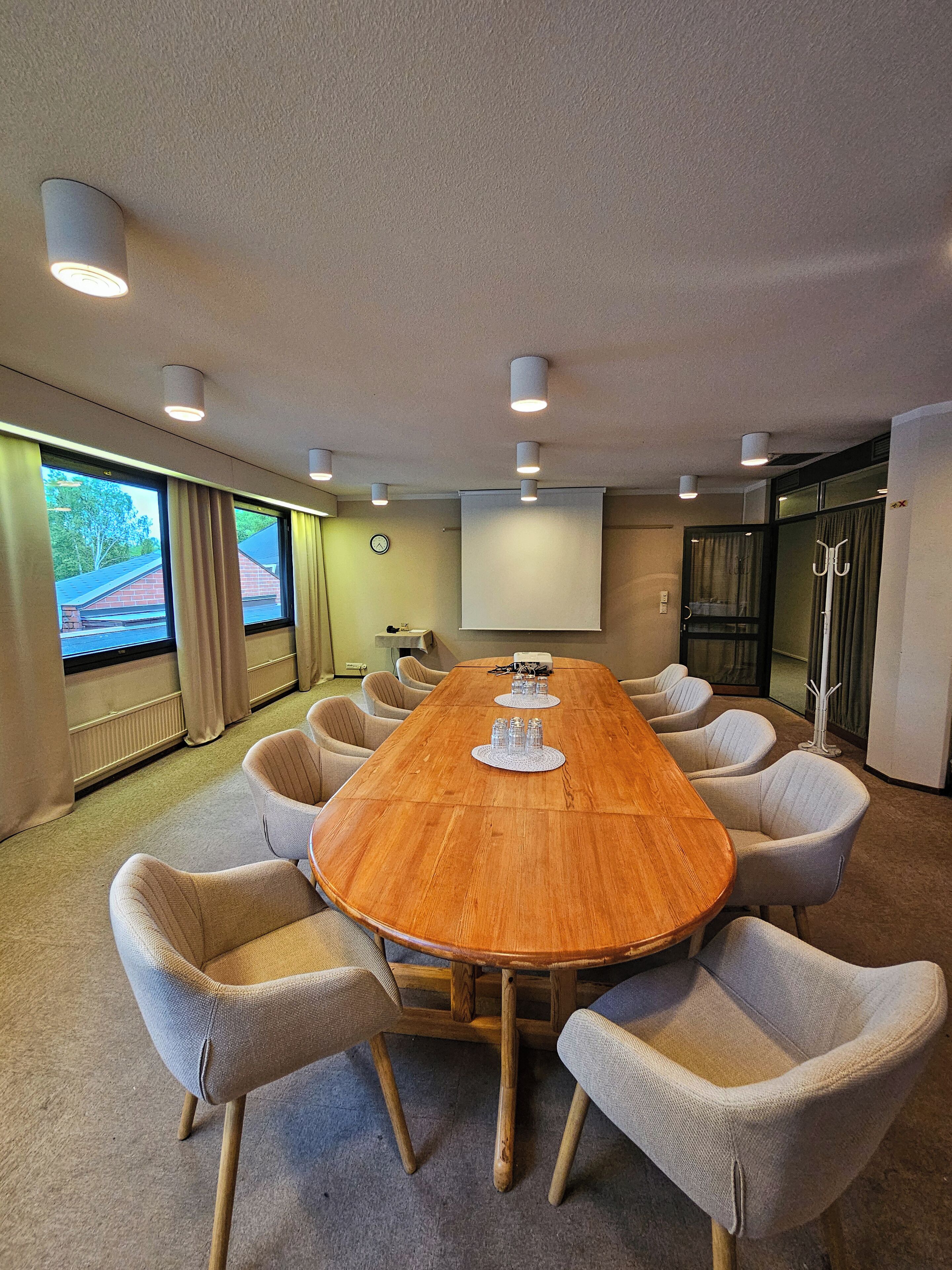 Un salón de conferencias con una mesa de madera, sillas color beige, una pantalla de proyector y un reloj en la pared.