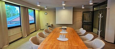 Un salón de conferencias con una mesa de madera, sillas color beige, una pantalla de proyector y un reloj en la pared.