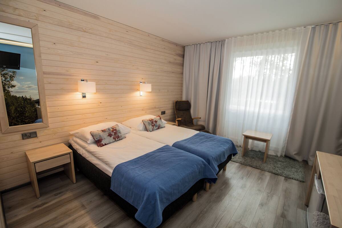 Quarto de hotel com duas camas, uma mesa pequena, uma cadeira, televisão na parede e janela com cortinas.